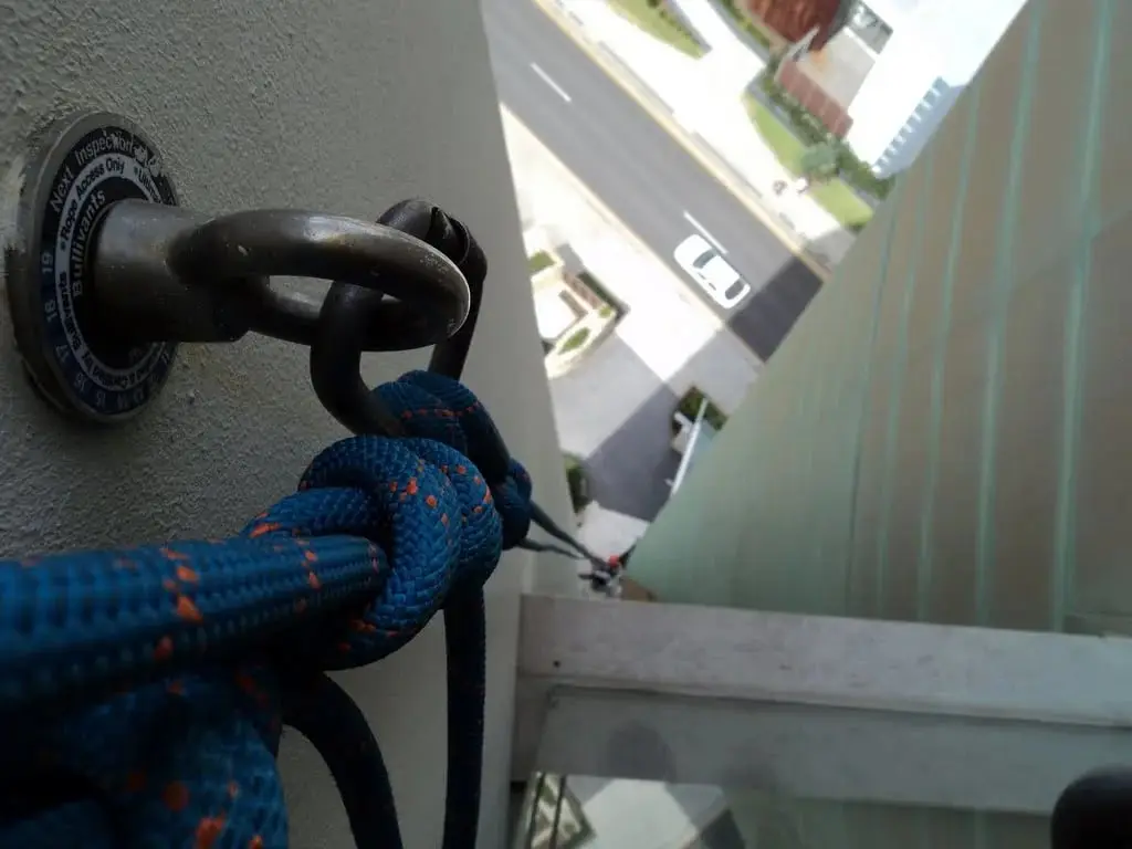 Blue rope tied off to a wall-mounted anchor eye at the top of a high-rise, with the road and parked cars far below