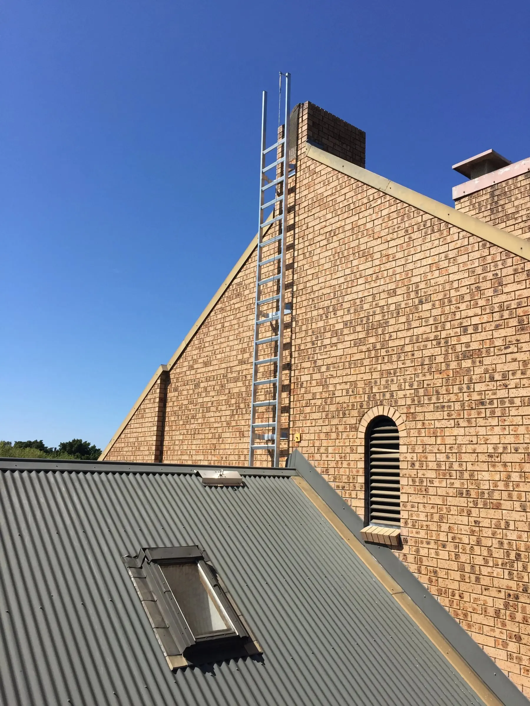New aluminium access ladder fixed up a brick gable wall, above a grey metal roof with a skylight