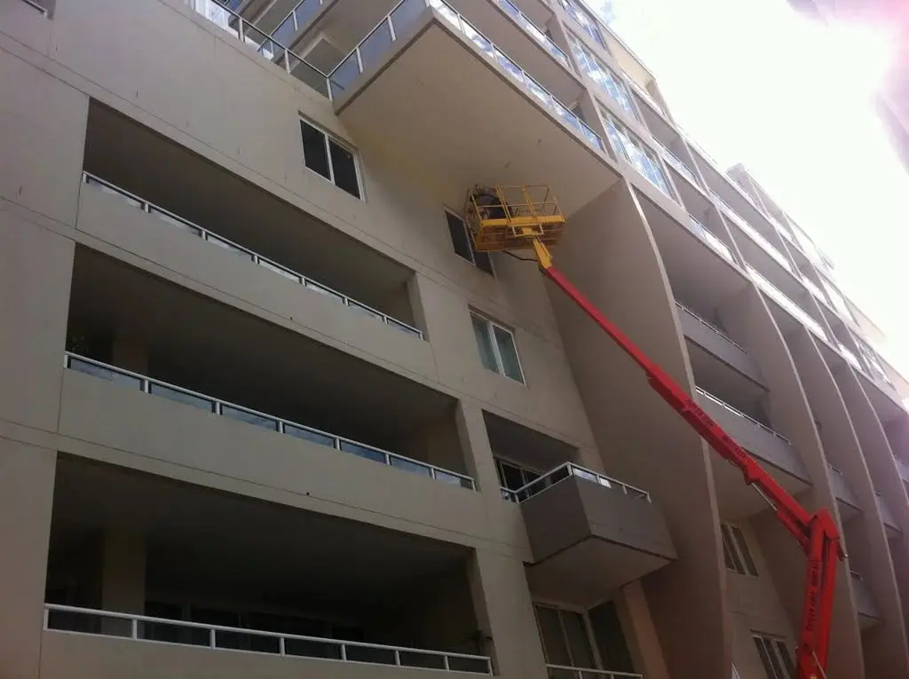 Boom lift with a red arm raising its yellow basket to the underside of a balcony on a multi-storey apartment building