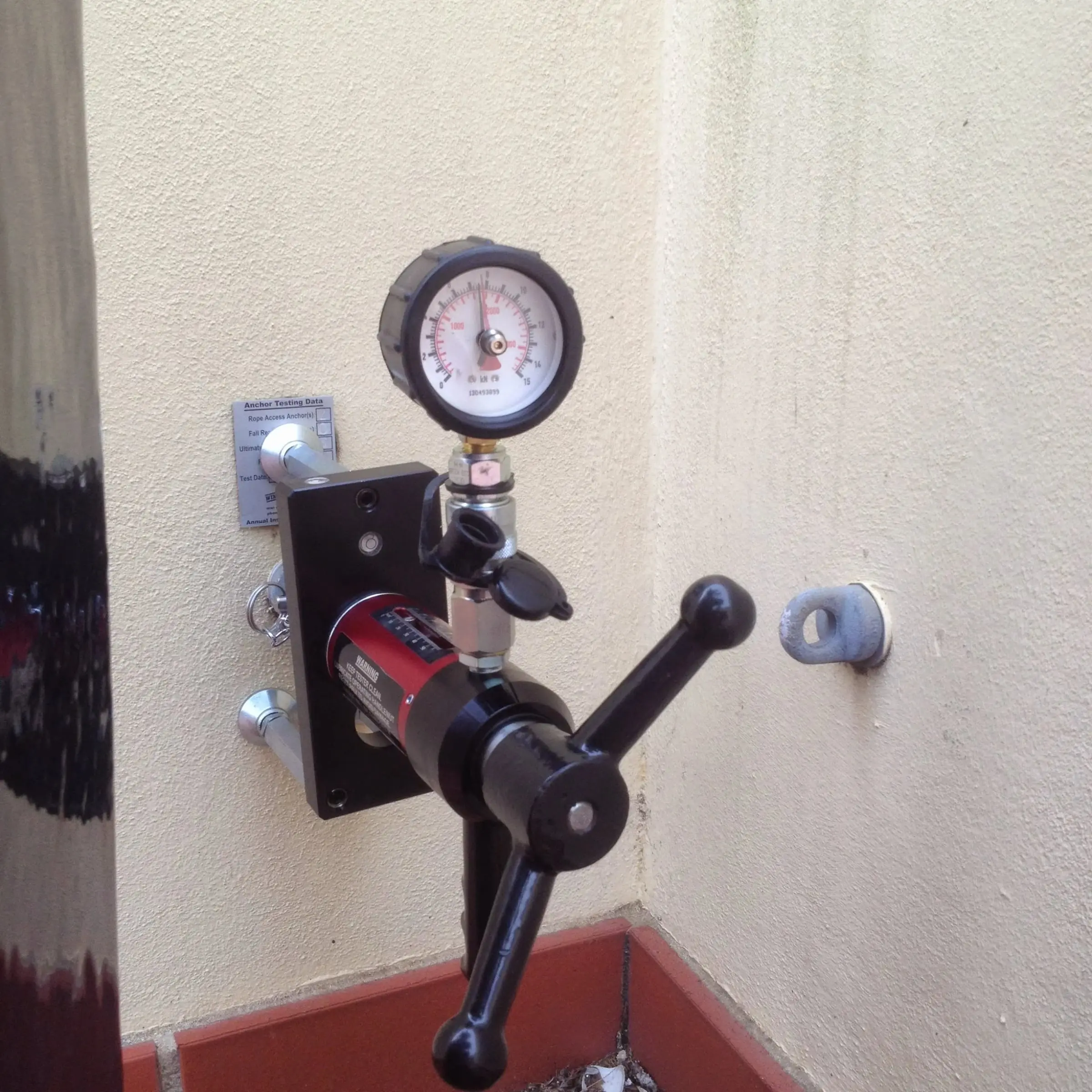 Hydraulic pull tester with a pressure gauge fitted to a wall anchor, below an anchor testing data plaque, with a second anchor eye on the wall beside it