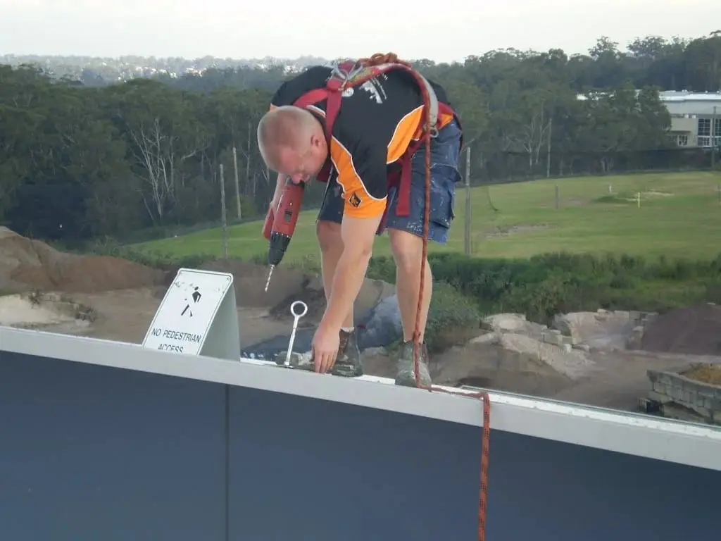 Technician in a harness drilling into a roof parapet beside a newly fitted anchor eye and a "No pedestrian access" sign