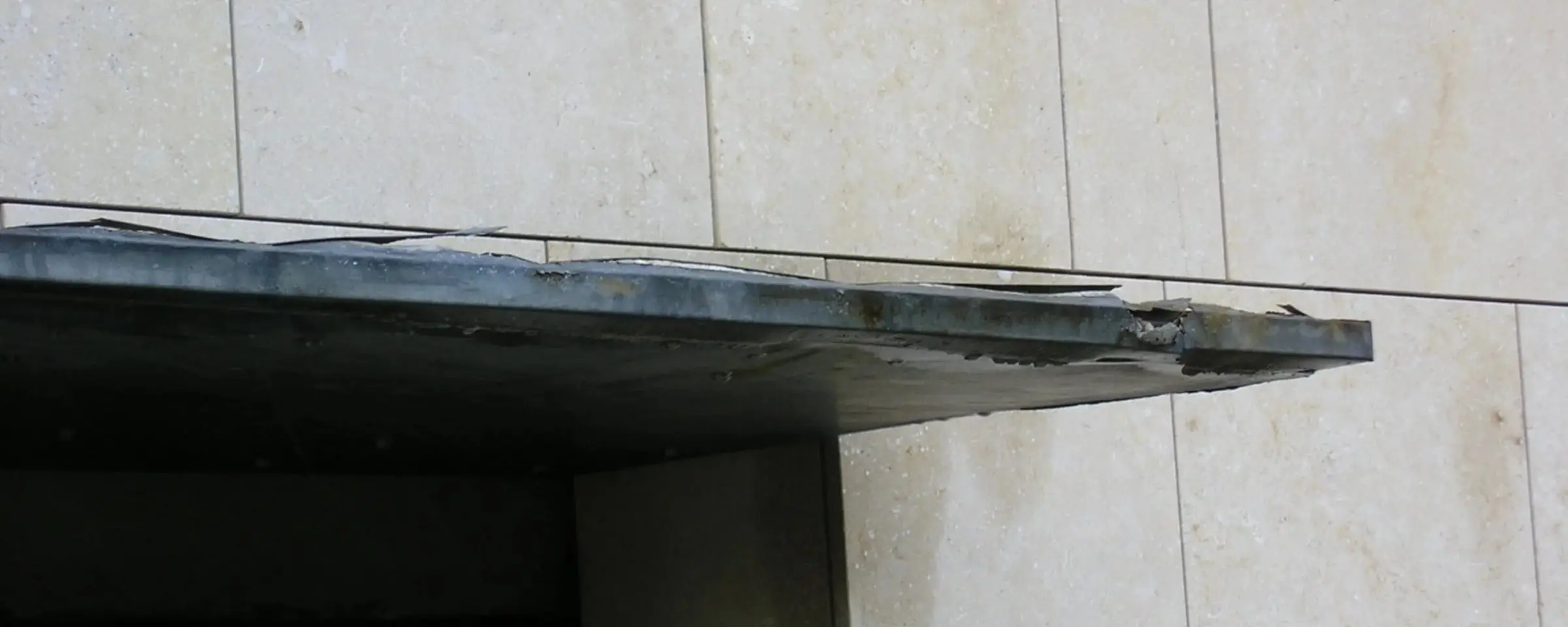 Corroded metal sunshade over a window on a pale stone-panelled wall, its front edge damaged and stained, before replacement