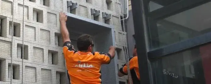 Fitting bird netting, Rope access technicians in orange shirts fitting mesh netting over a patterned block facade beside a glass lift shaft.