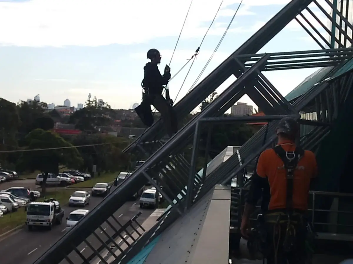 Two OneVision technicians, one on ropes, working on a steel awning frame, Gateway, Maroubra