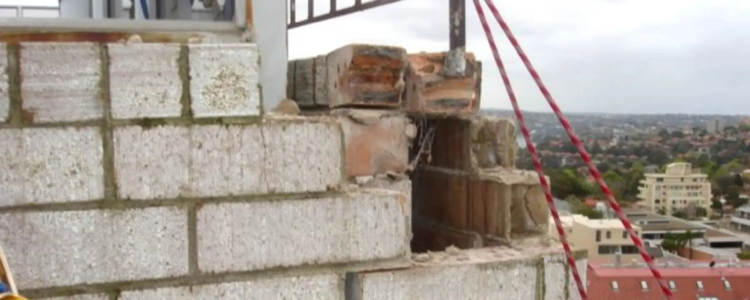 Top of a brick parapet with broken and missing bricks, a red access rope running past and suburbs in the distance