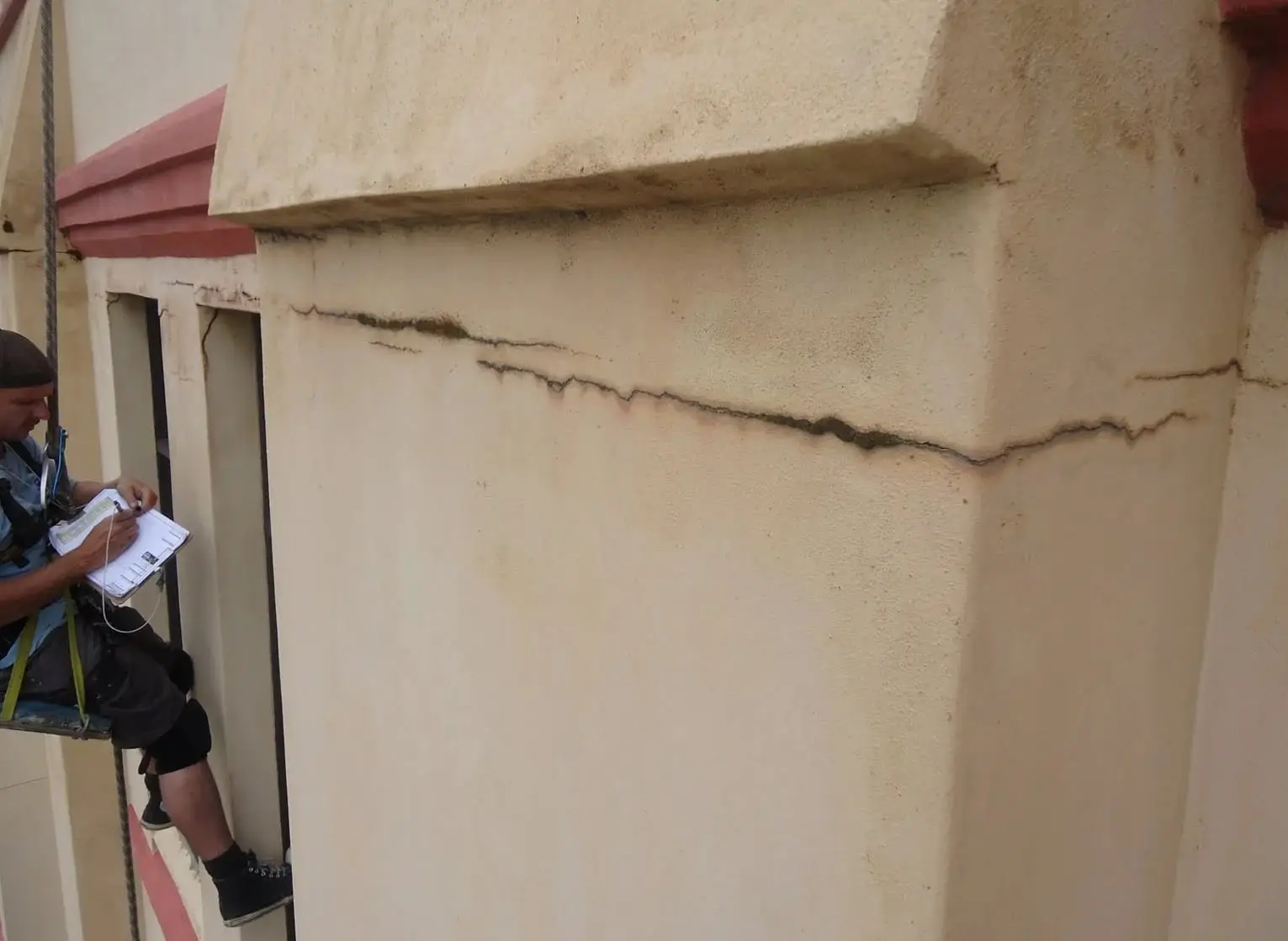 Rope access technician in a harness writing on a clipboard beside a long horizontal crack in the cream render of a building facade