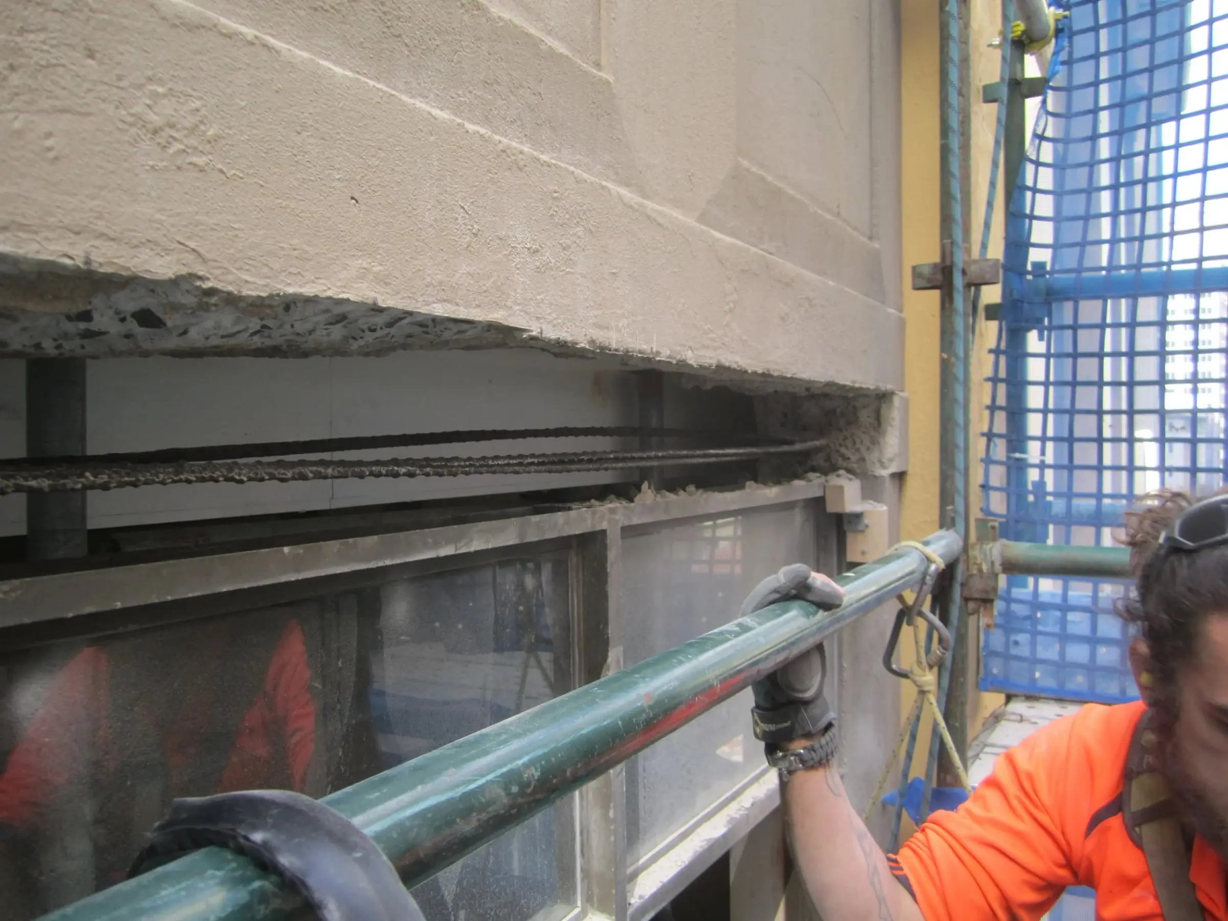 Technician holding a scaffold rail beside a concrete beam cut back to expose its reinforcing bars, above a shopfront window