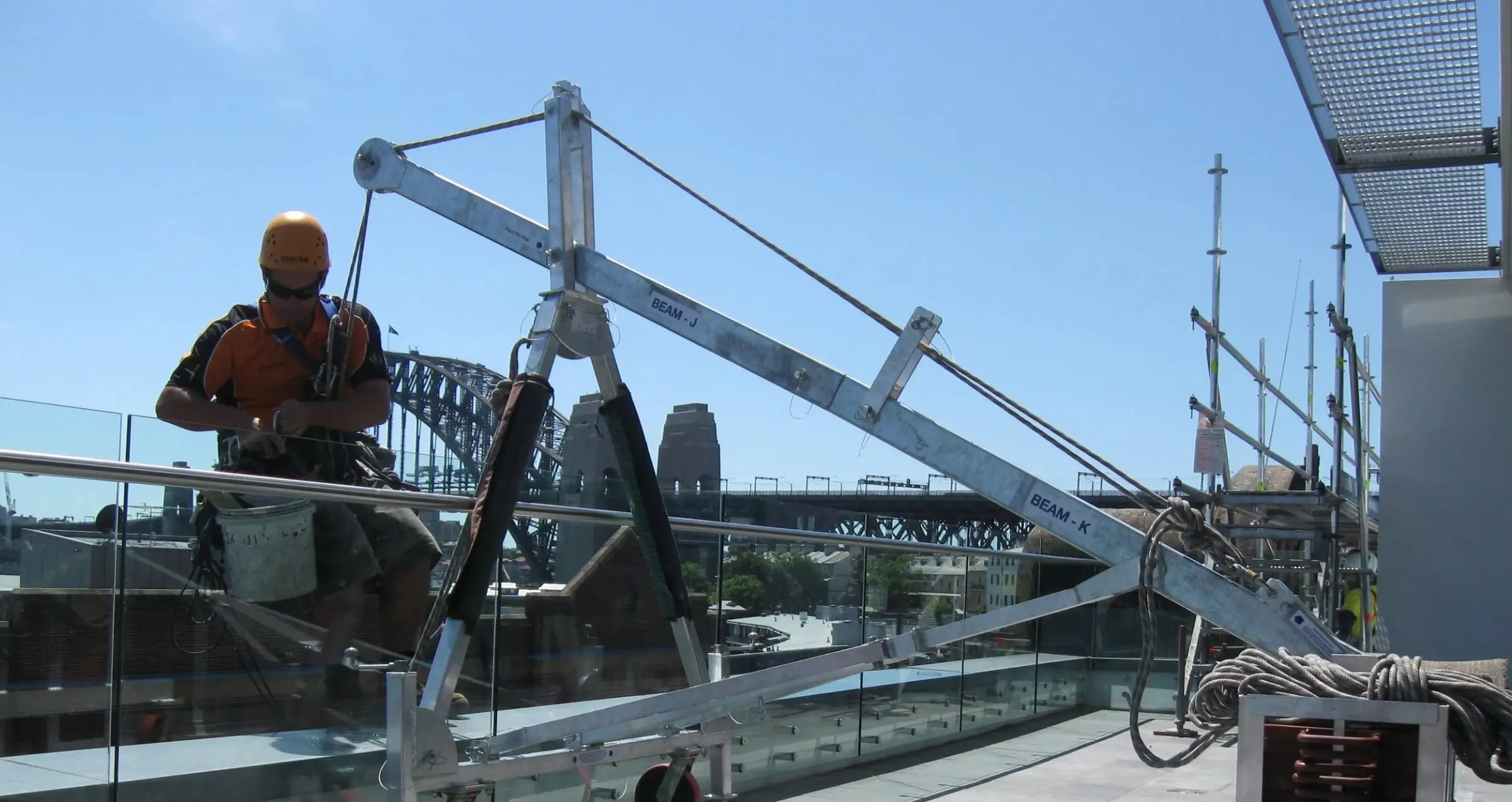 Technician in a helmet and harness beside an aluminium davit arm on a glass-balustraded rooftop, with the Sydney Harbour Bridge behind