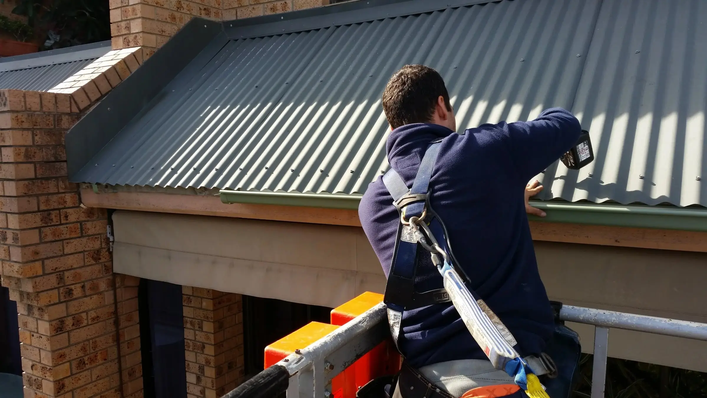 Technician in a safety harness on an elevated work platform fixing a new gutter along the edge of a corrugated metal roof on a brick building