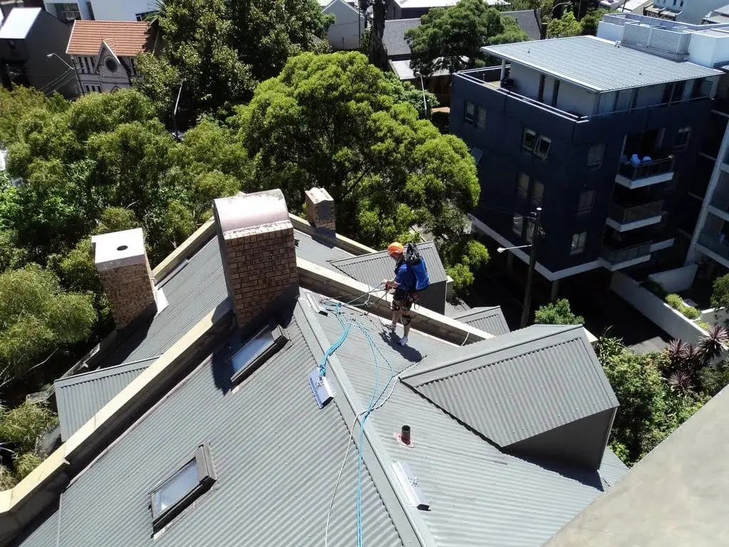 Roof access set up, A technician on a pitched metal roof with access ropes rigged across it, among apartment buildings.