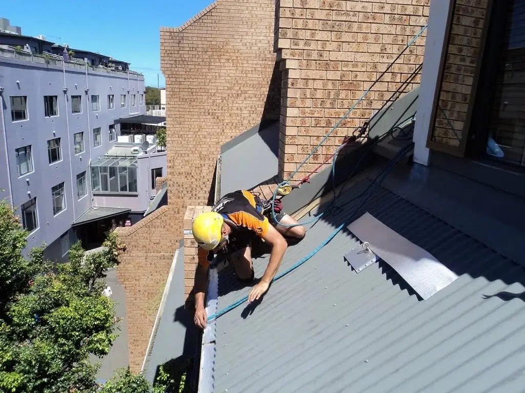 Working at the roof edge, A roped technician reaching into the gutter where a corrugated roof meets a brick wall.