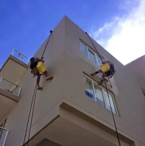 Two painters on the facade, Two rope access painters with paint buckets working down a rendered wall beside the windows.