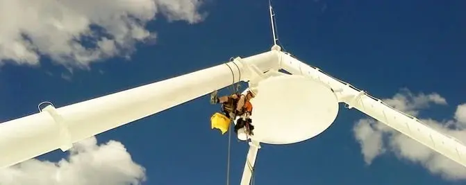 Painting a satellite dish, A roped painter hanging beside a large white dish on a steel frame, against a blue sky.