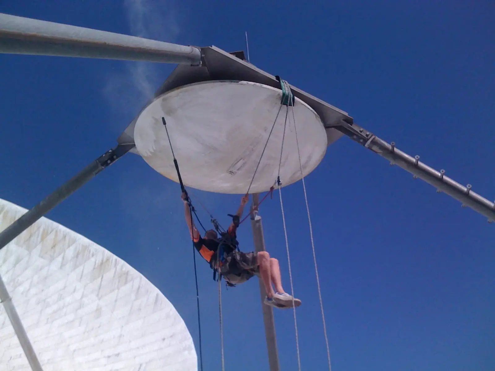 Rope access technician hanging beneath a large round dish on a steel frame, washing its underside with a long spray lance against a blue sky