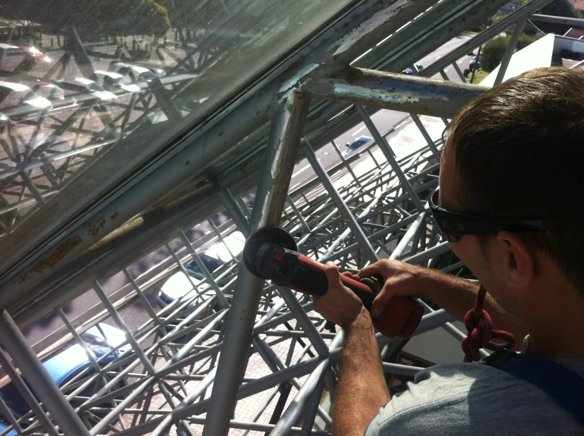Technician in safety glasses grinding a steel frame member on a lattice structure beneath glass panels