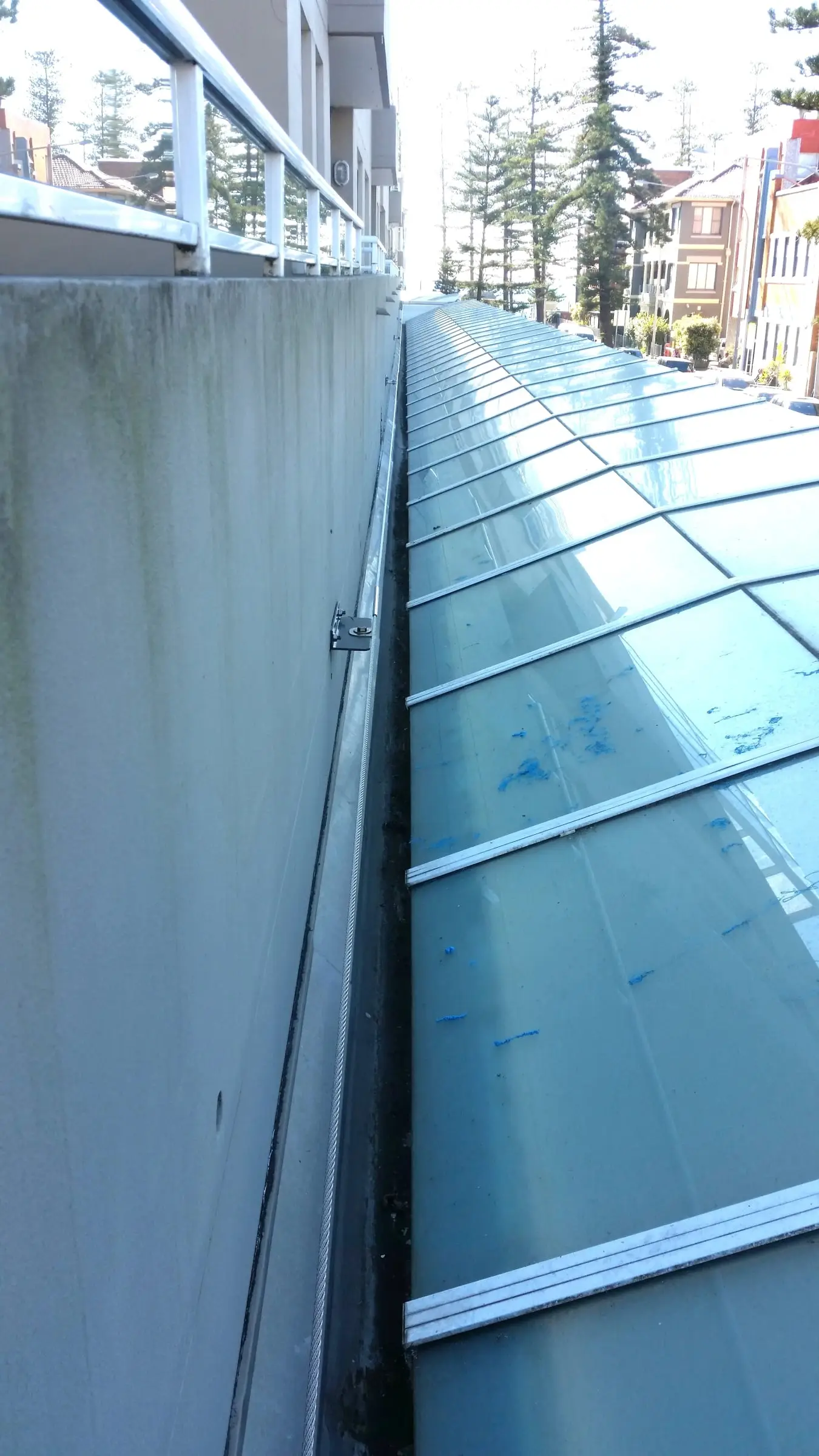 Steel cable line fixed with brackets along a rendered wall, beside a long sloping glass roof