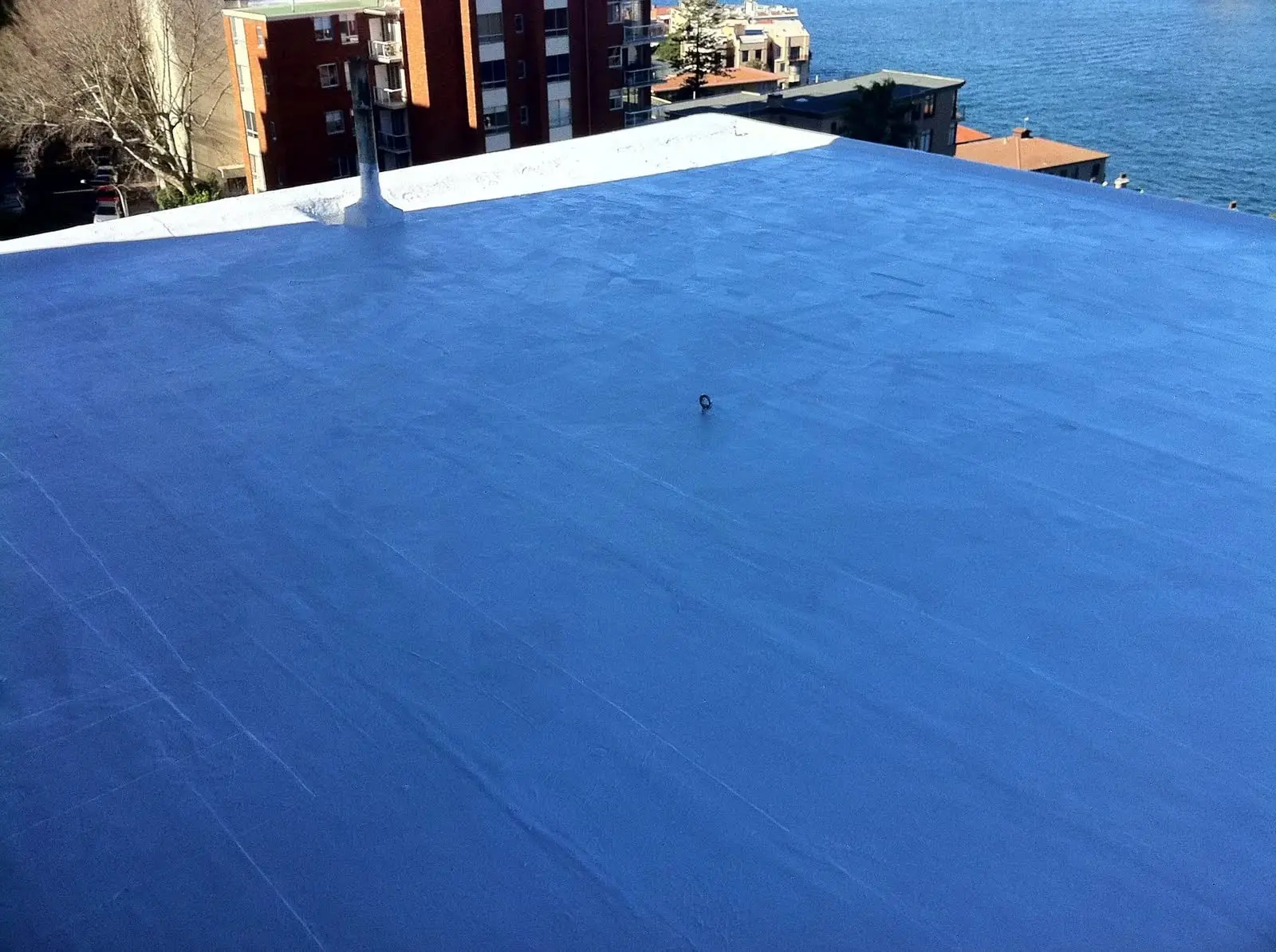 A flat rooftop freshly coated in a smooth blue waterproofing membrane, with apartment buildings and the harbour beyond