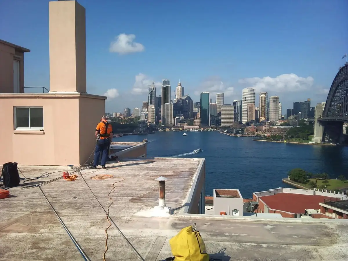Before, Kirribilli roof, A technician on a weathered flat roof overlooking the harbour and the city skyline, before waterproofing.