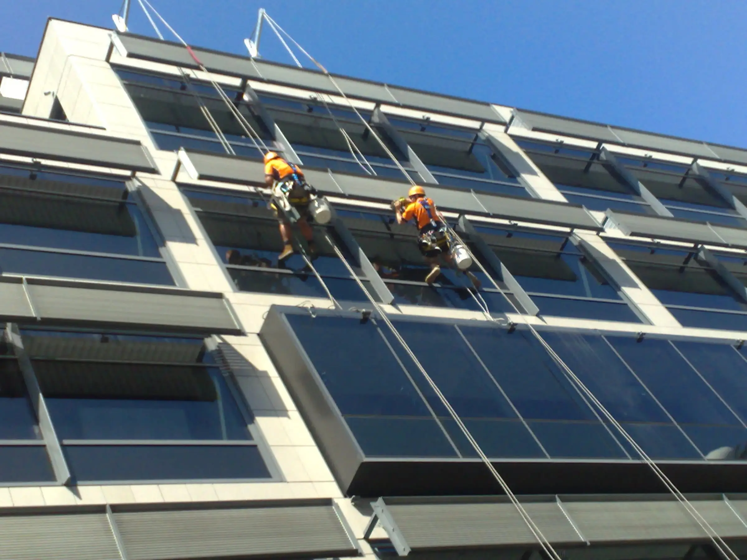Two rope access technicians in orange shirts and helmets cleaning the windows of an office building, hanging from ropes rigged off the roof