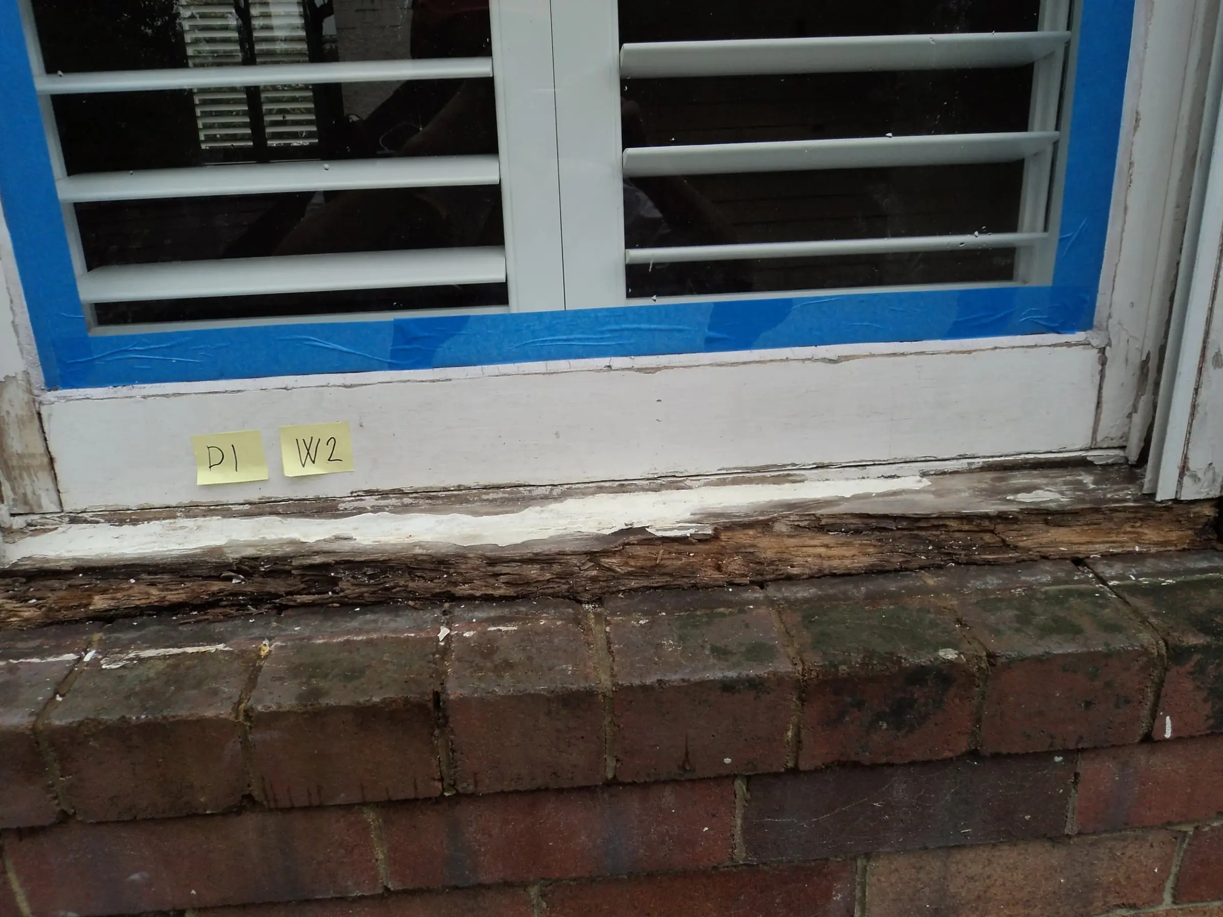 Timber window with its glass masked in blue tape above a weathered sill with flaking paint and rotted timber, before restoration
