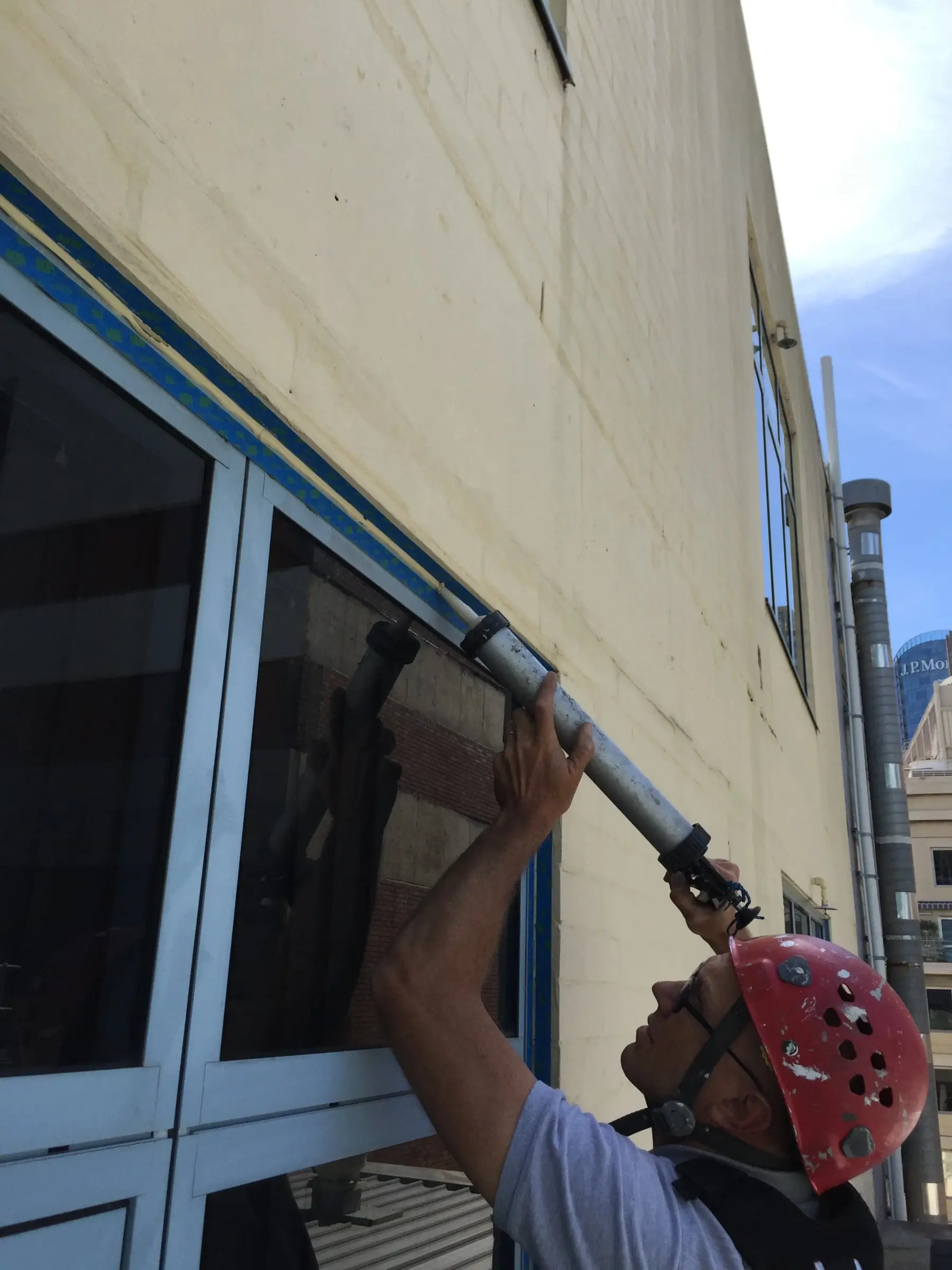 Resealing a window frame, A roped technician in a red helmet applying sealant along the top of a window frame, taped off in blue.