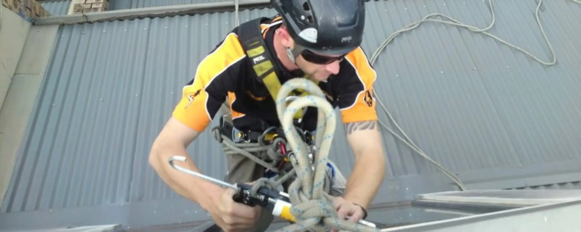 Rope access technician in a helmet and harness running a sealant gun along a metal frame set into a corrugated metal roof