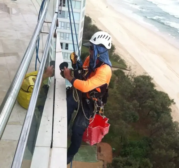 Beachfront high-rise, A technician on ropes working on a balcony glass balustrade high above a beach.