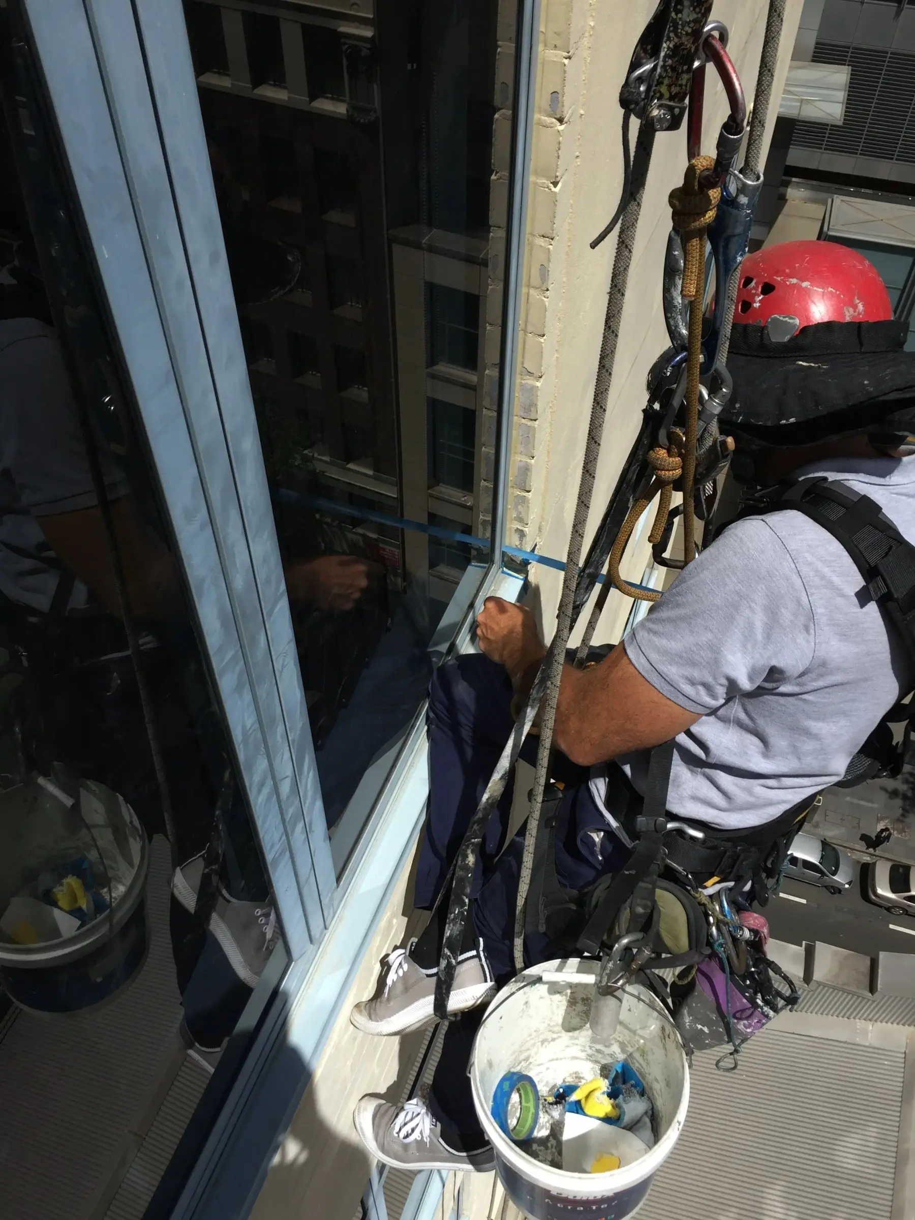 Sealing from a rope, A technician in a seat harness working on a window frame, with his gear and a bucket hanging beside him.