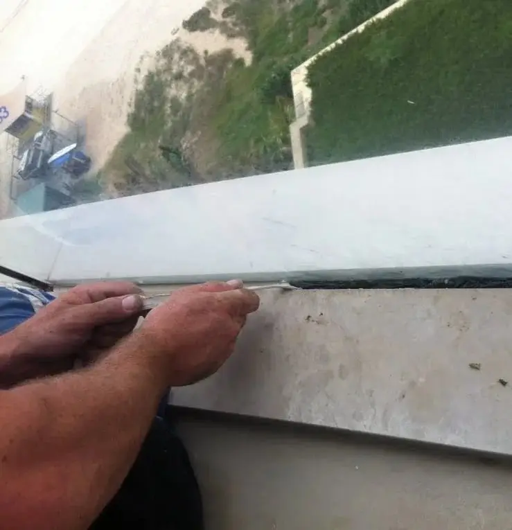 Tooling the sealant, A hand smoothing a fresh bead of sealant along a stone ledge beneath the glass.