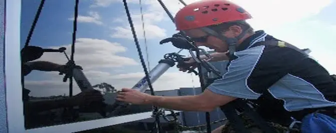 Working at the window, A roped technician in a red helmet sealing around a window, with the sky reflected in the glass.